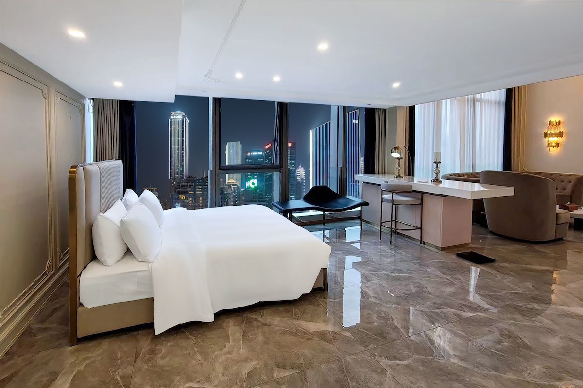 A hotel room with a bed, a desk, a chair, and a view of a cityscape.