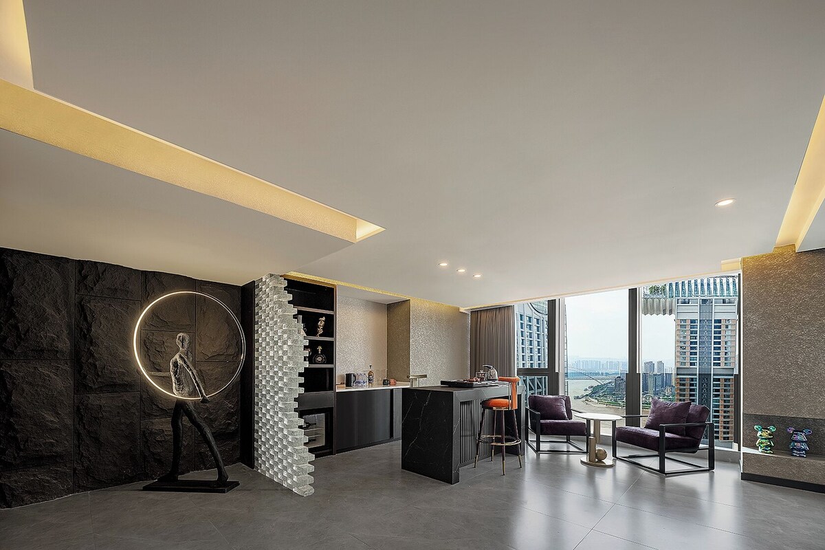 A modern hotel room with a marble wall, a circular sculpture, a bar area, and a view of the city.