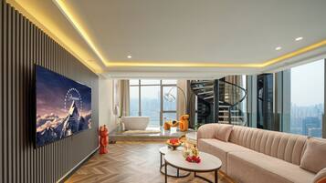 Ein modernes Wohnzimmer mit großem Flachbildfernseher, einer Couch, einem Couchtisch und Blick auf eine Stadtlandschaft durch hohe Fenster.