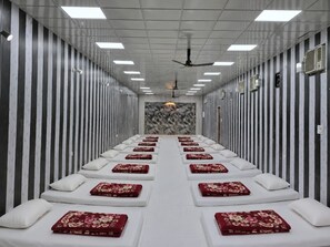 Una habitación con una hilera larga de camas alineadas a lo largo de un pasillo central, cada cama con almohada y una manta roja estampada.