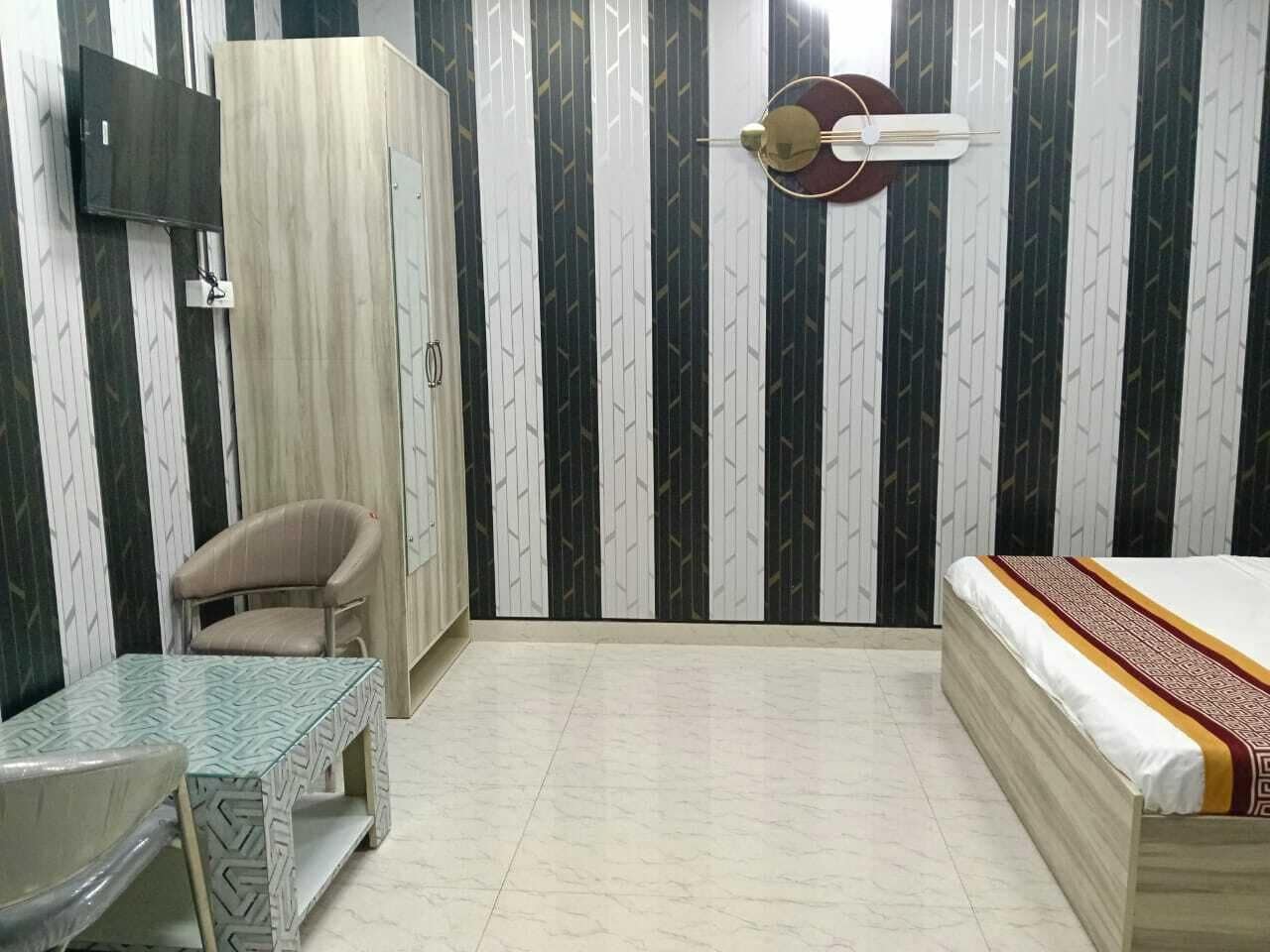 A bedroom with a bed, a chair, a table, a wardrobe, and a wall-mounted TV.
