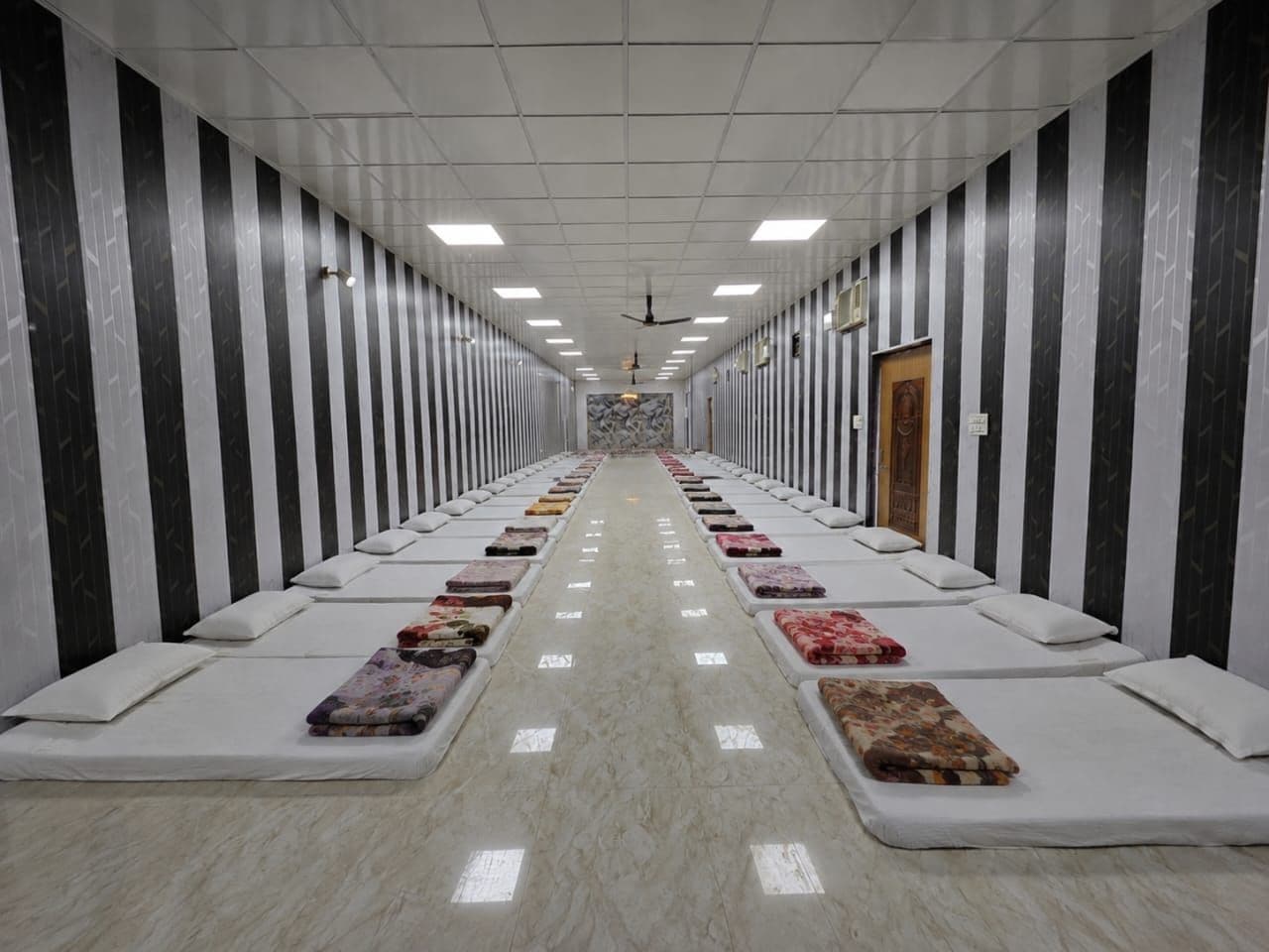 A hotel corridor with rows of beds, a ceiling fan, and a door at the end.