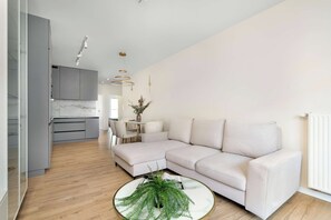 A modern living room with a light-colored sofa, a round coffee table with a plant, and a kitchen area with grey cabinets and pendant lighting. - Spacious Apartment with a balcony | Close to the Airport (Poznań)