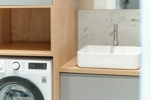 A washing machine with a digital display, a wooden shelf, and a white sink with a marble countertop. - Spacious Apartment with a balcony | Close to the Airport (Poznań)