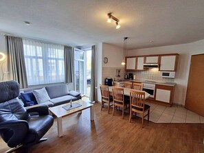 Ein modernes Wohnzimmer mit einer Couch, einem Couchtisch und einem Essbereich mit Stühlen. Im Hintergrund ist eine Küche mit Holzausstattung und Geräten zu sehen.