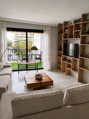 Ein modernes Wohnzimmer mit einer Couch, einem Couchtisch, einem Fernseher und einem Balkon mit Blick auf Gebäude und Grünflächen.