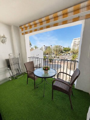 Ein Balkon mit Kunstgrashalm, zwei Korbstühle, einem Glastisch mit Blumenarrangement und einem Metallgeländer.
