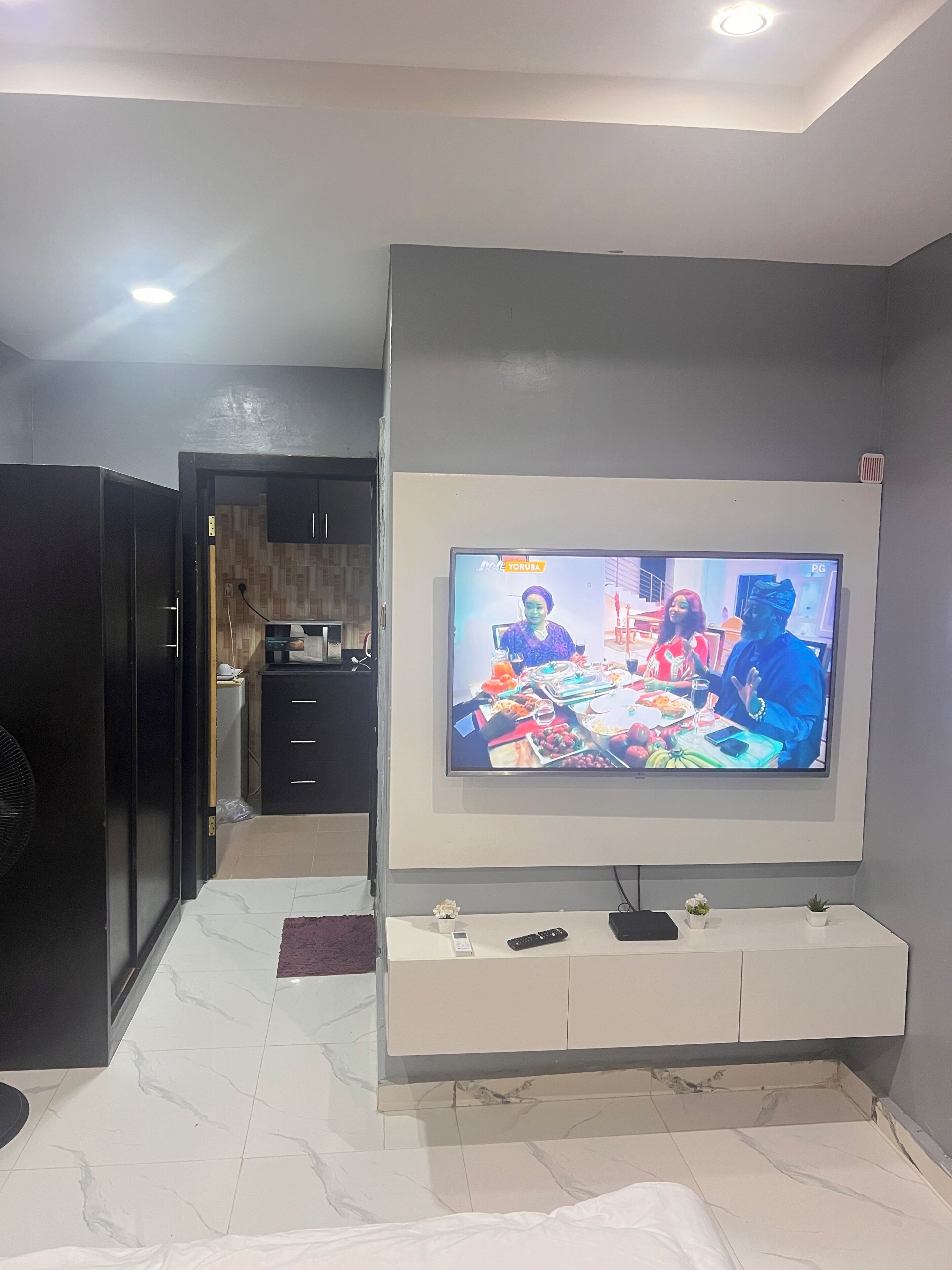 Una habitación con una televisión montada en la pared, un mueble blanco debajo y piso de baldosas.