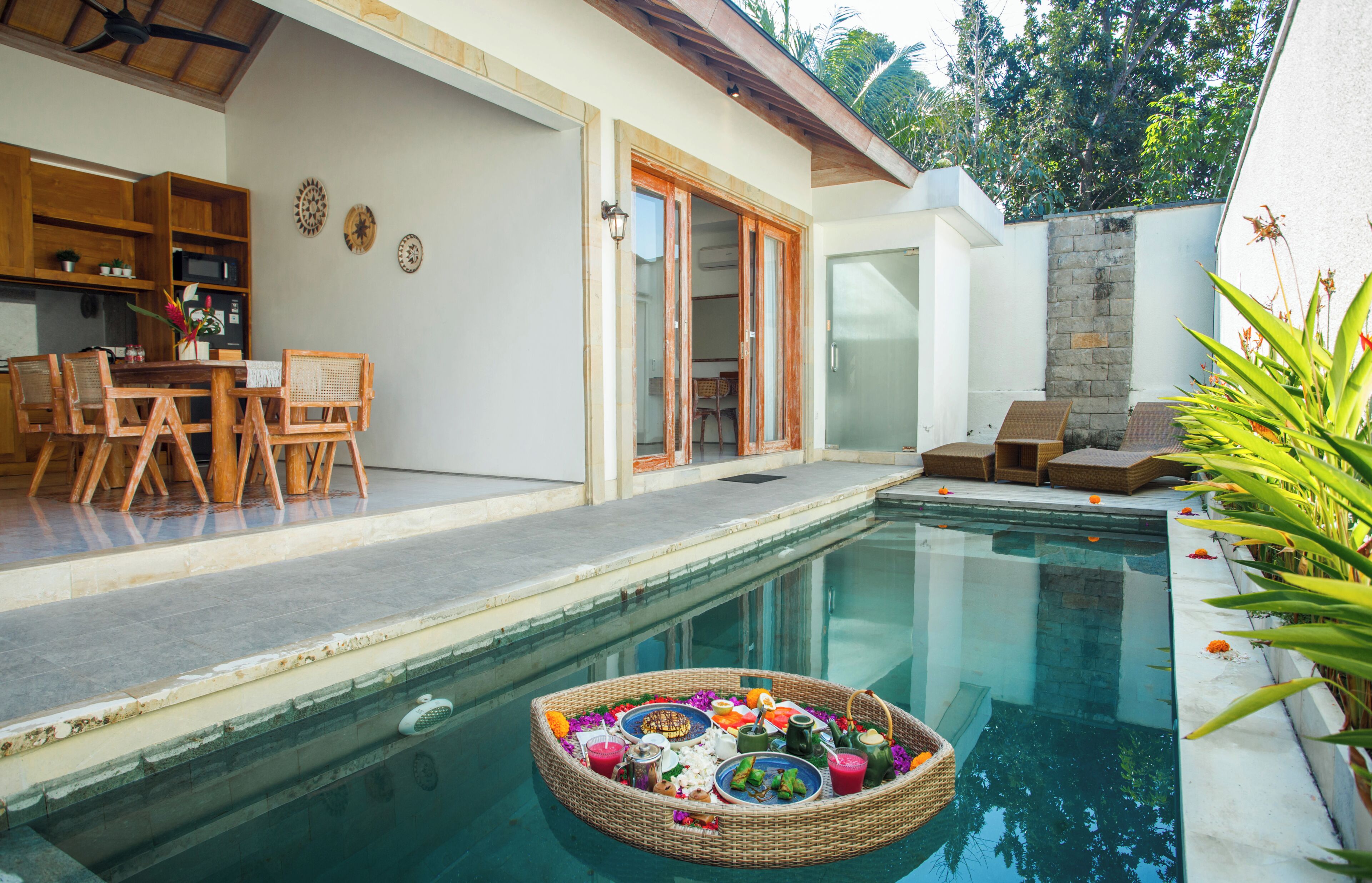 Una piscina con una cesta flotante de comida y bebidas, un área de comedor con sillas y una mesa, y un espacio de estar techado al aire libre.