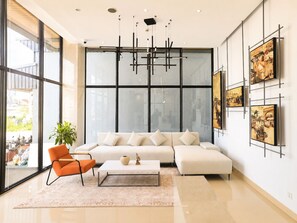 A modern living room with a white sofa, orange chair, and a coffee table. There are large windows and artwork on the walls. - One Studio Suite by PSH (Ou Chrov)