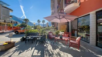Un espace extérieur aménagé avec des tables et des chaises, un grand parasol, et un bâtiment doté de grandes fenêtres et de balcons.