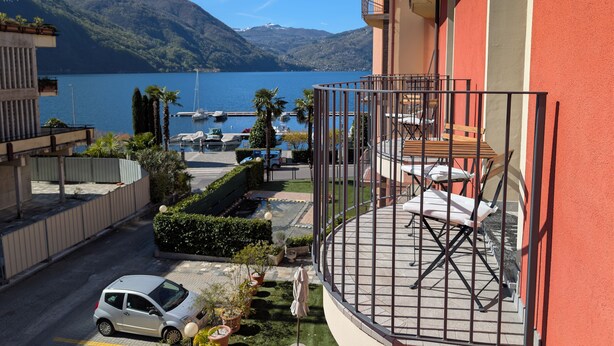 Un balcón con vistas a un lago y montañas, un coche estacionado y un edificio con fachada roja.