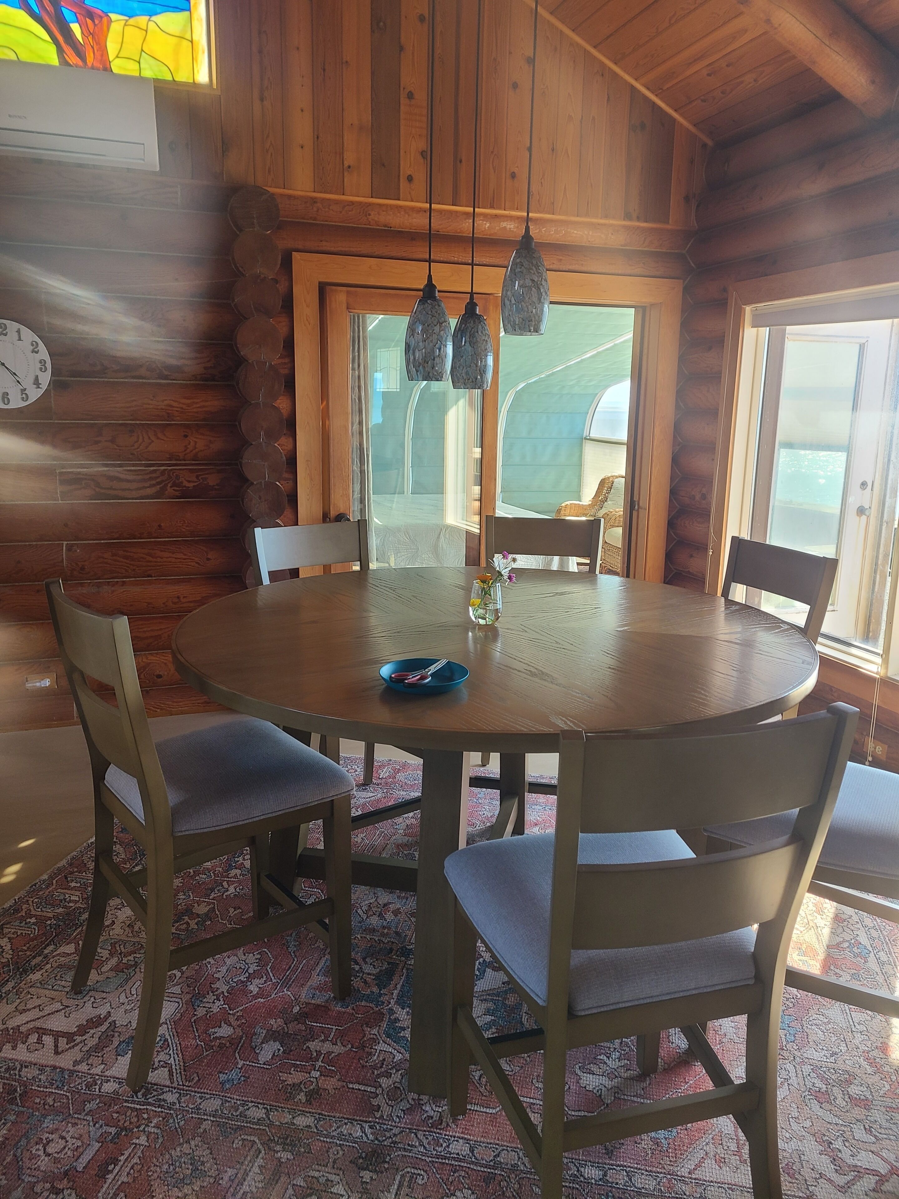 Un interior de madera con una mesa redonda, sillas, un reloj y vistas a través de una ventana.