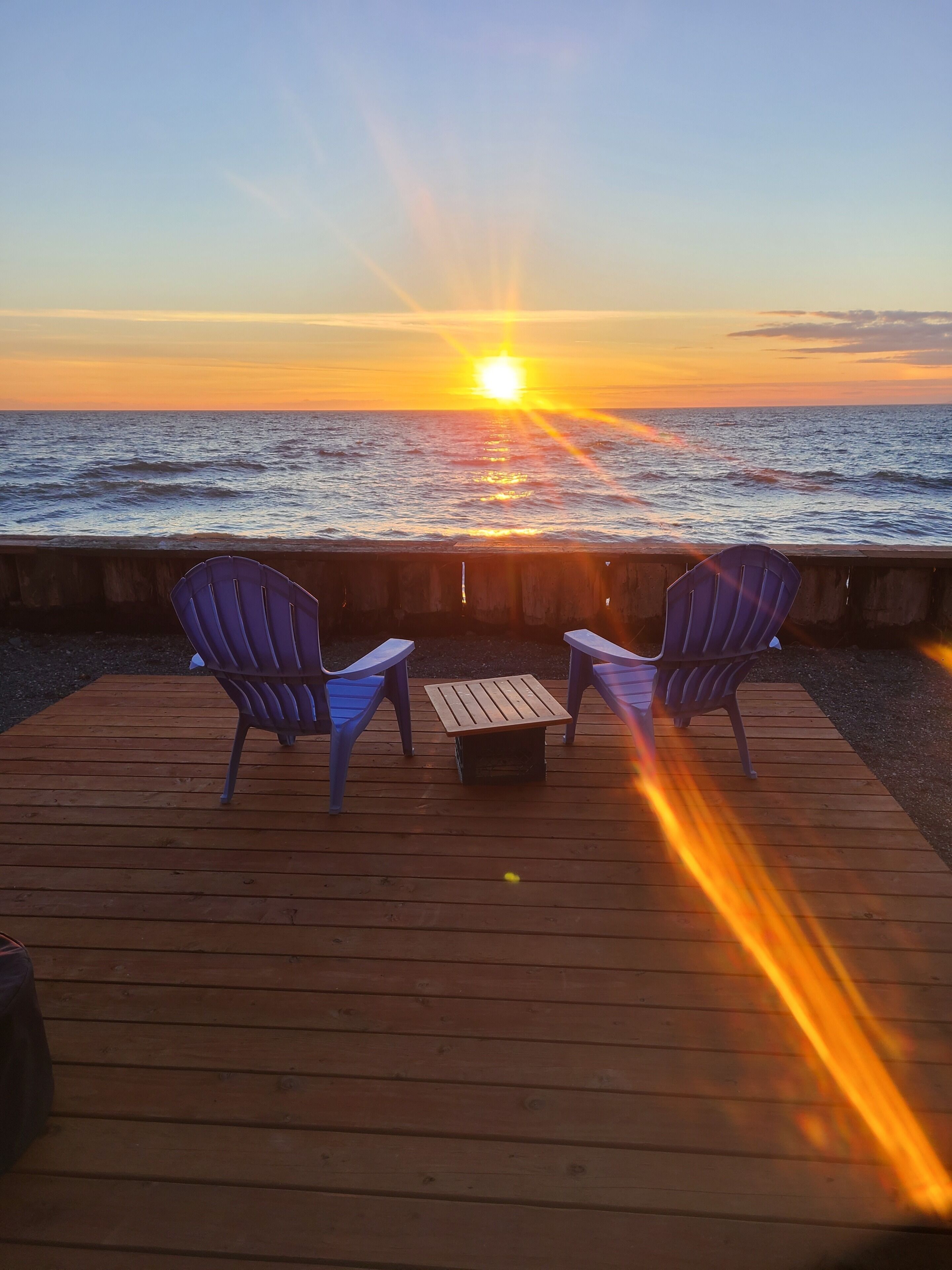 En træterrasse med to stole og et bord, med udsigt til en solnedgang over havet.