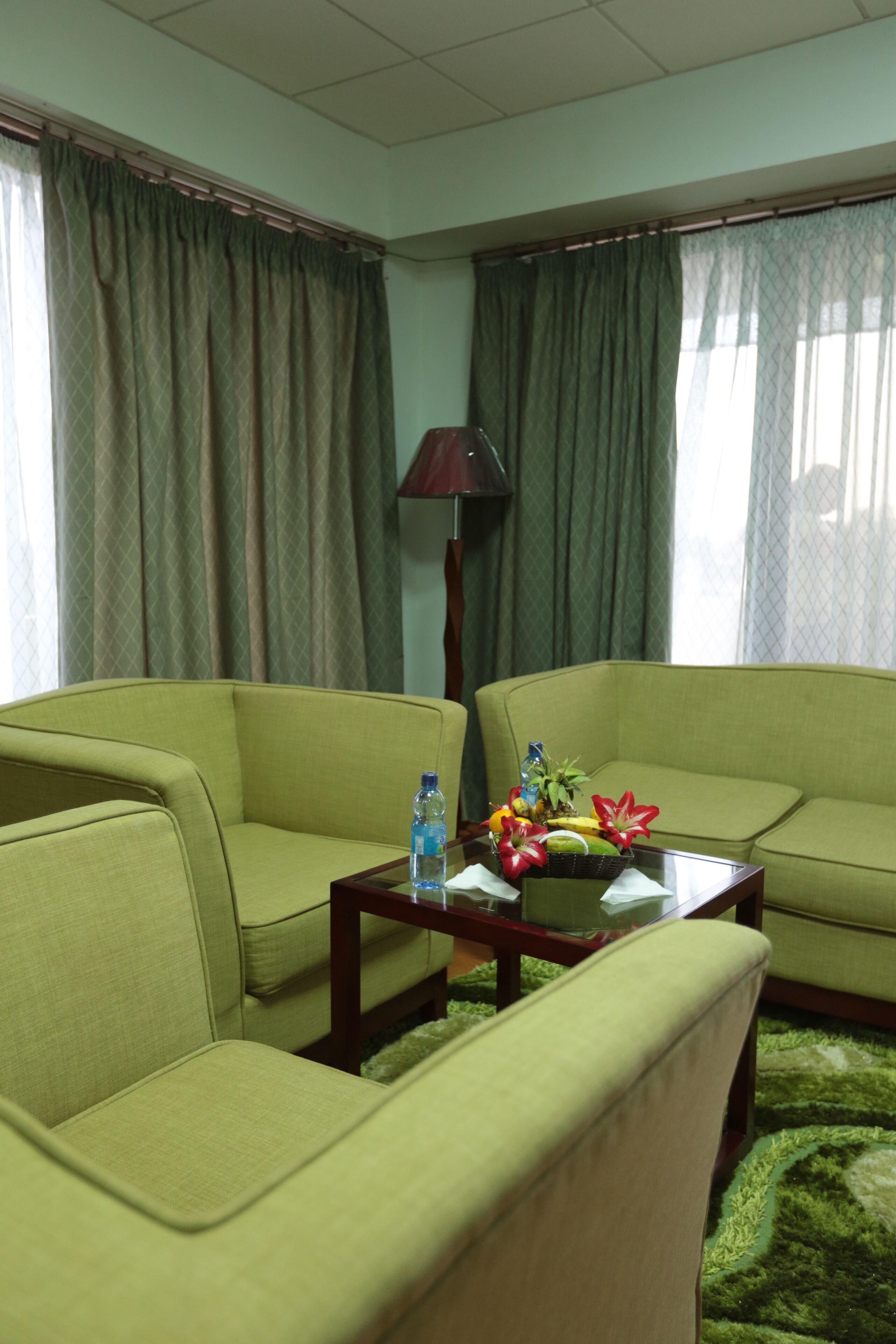 Quarto com dois sofás verdes, uma mesa de centro de vidro com uma cesta de frutas, uma luminária e vista para o exterior através de cortinas.