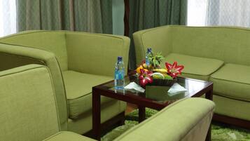 Quarto com dois sofás verdes, uma mesa de centro de vidro com uma cesta de frutas, uma luminária e vista para o exterior através de cortinas.