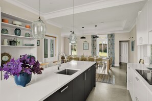 A modern kitchen with a central island, black cabinets, and a white countertop. There are hanging pendant lights, a dining area with wooden chairs, and a view of the outdoors through a window. - Russell Bank is a beautiful house with amazing sea views in the heart of Elie. (Elie and Earlsferry)