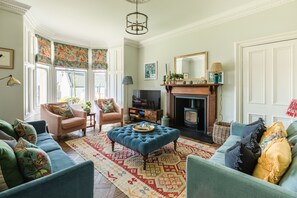 A traditional living room with a fireplace, comfortable seating, and a large window with patterned curtains. - Russell Bank is a beautiful house with amazing sea views in the heart of Elie. (Elie and Earlsferry)