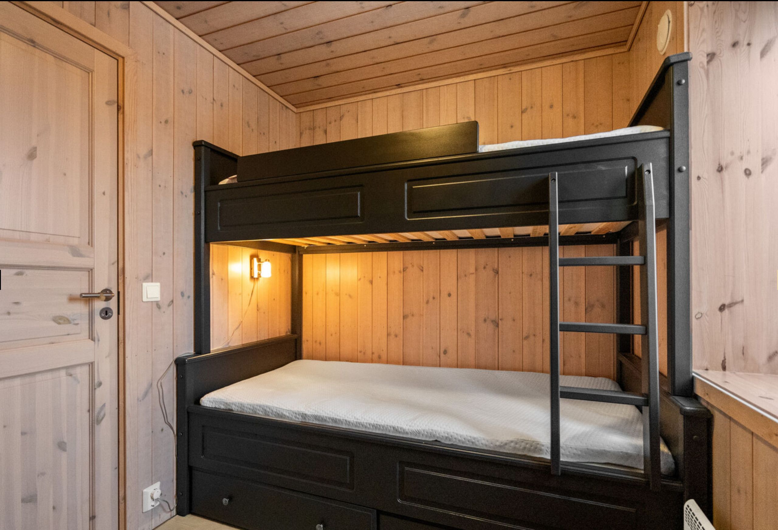 Una cama litera con escalera, una luz fijada en la pared y una puerta de madera.