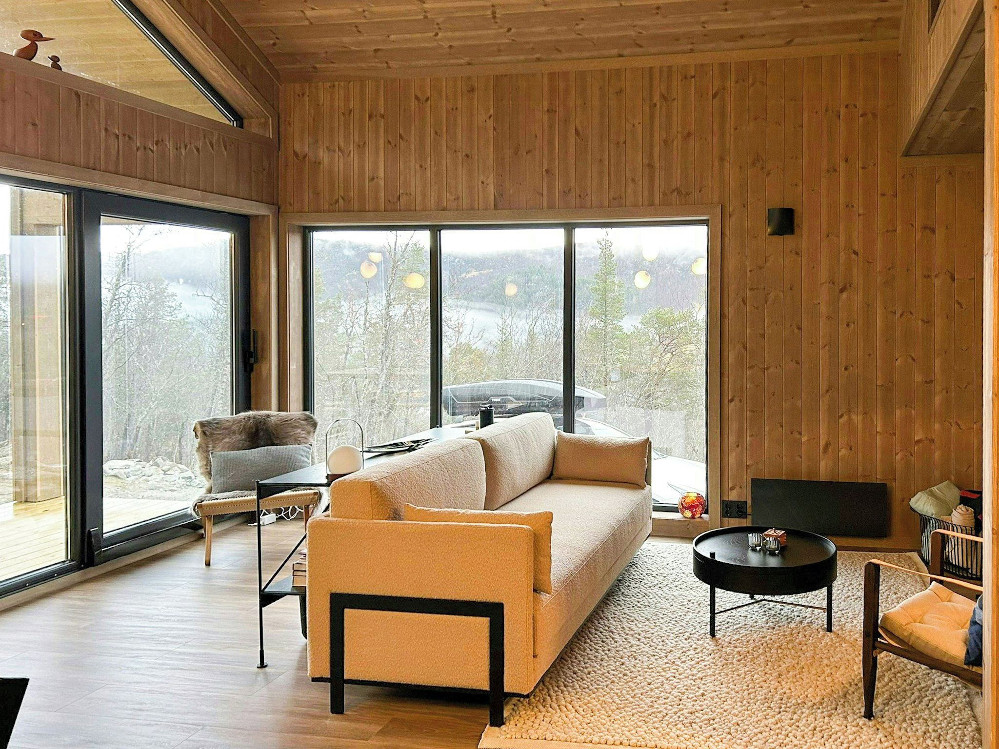 A modern living room with a sofa, armchair, and a TV. Large windows offer a view of trees and a lake.