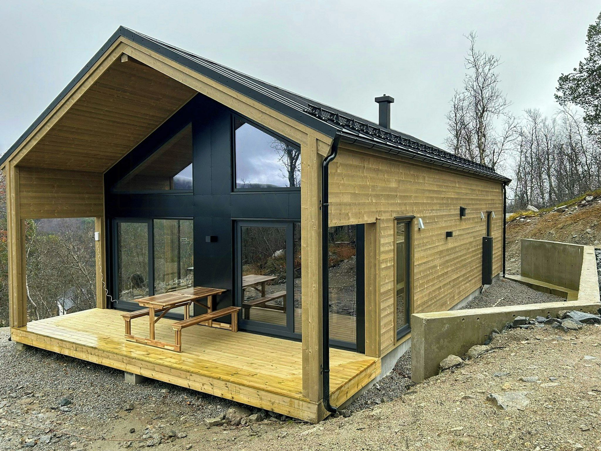 En modern stuga i trä med en stor veranda, ett picknickbord och en skorsten.