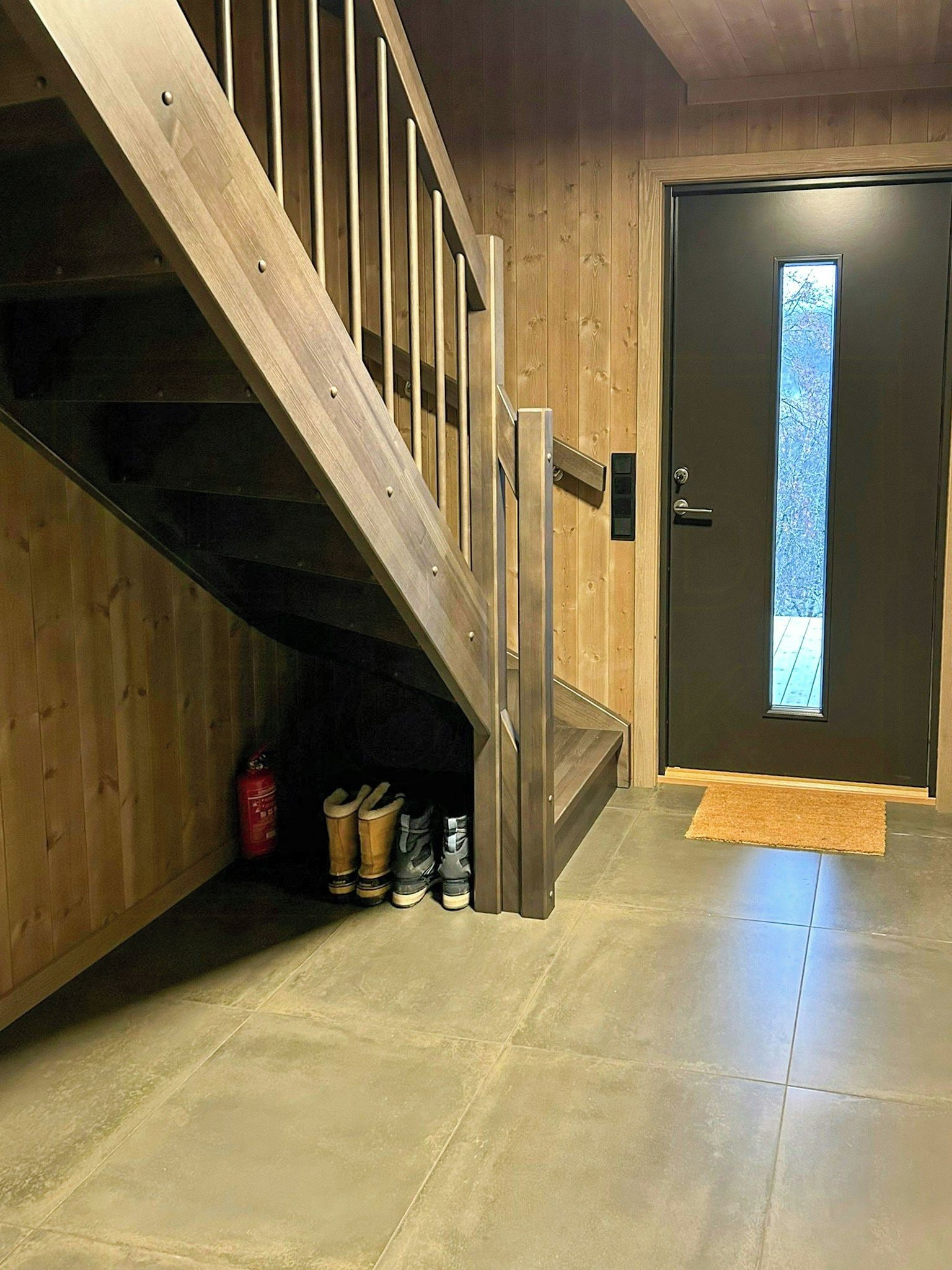 Eine Holztreppe führt zu einer unteren Ebene, eine Tür mit Fenster und ein Feuerlöscher stehen auf dem Boden.