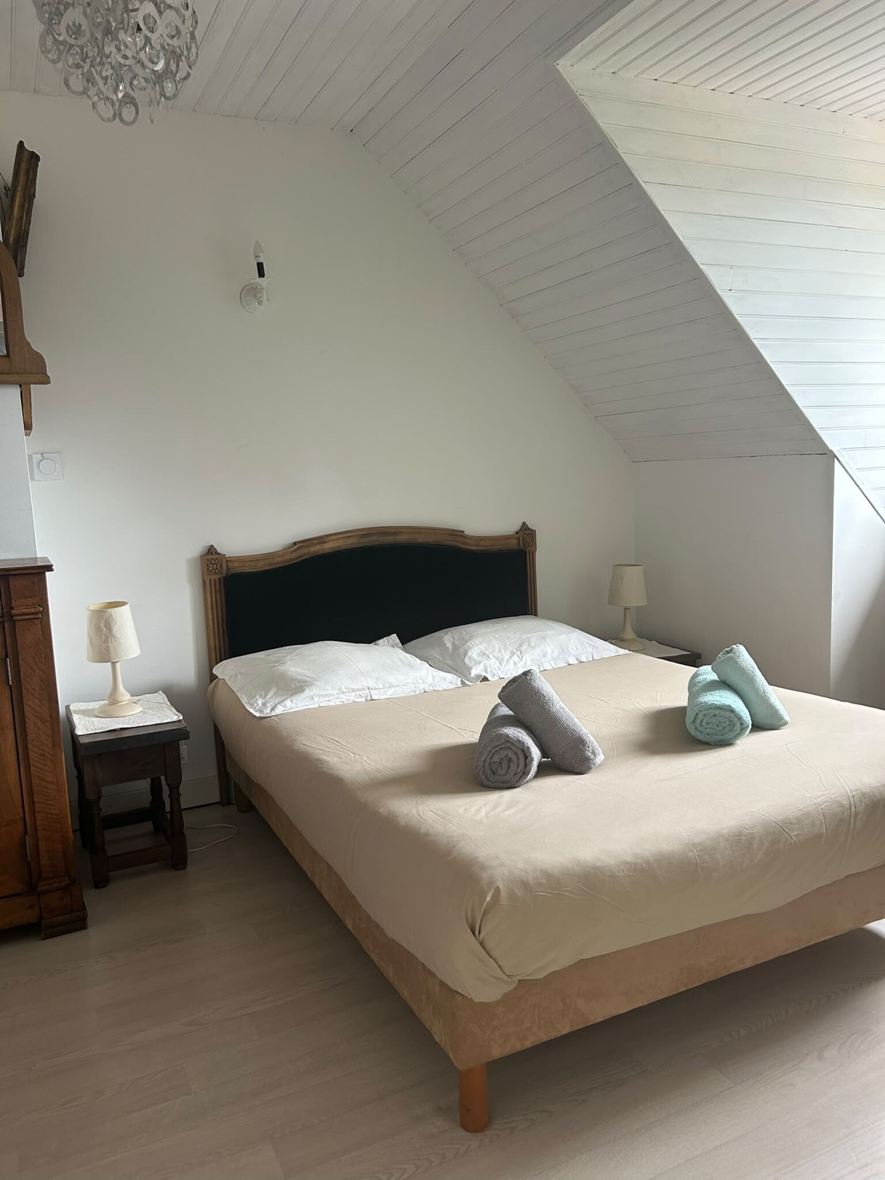 A bedroom with a sloped ceiling, a bed with white linens, two bedside lamps, and a wooden nightstand.