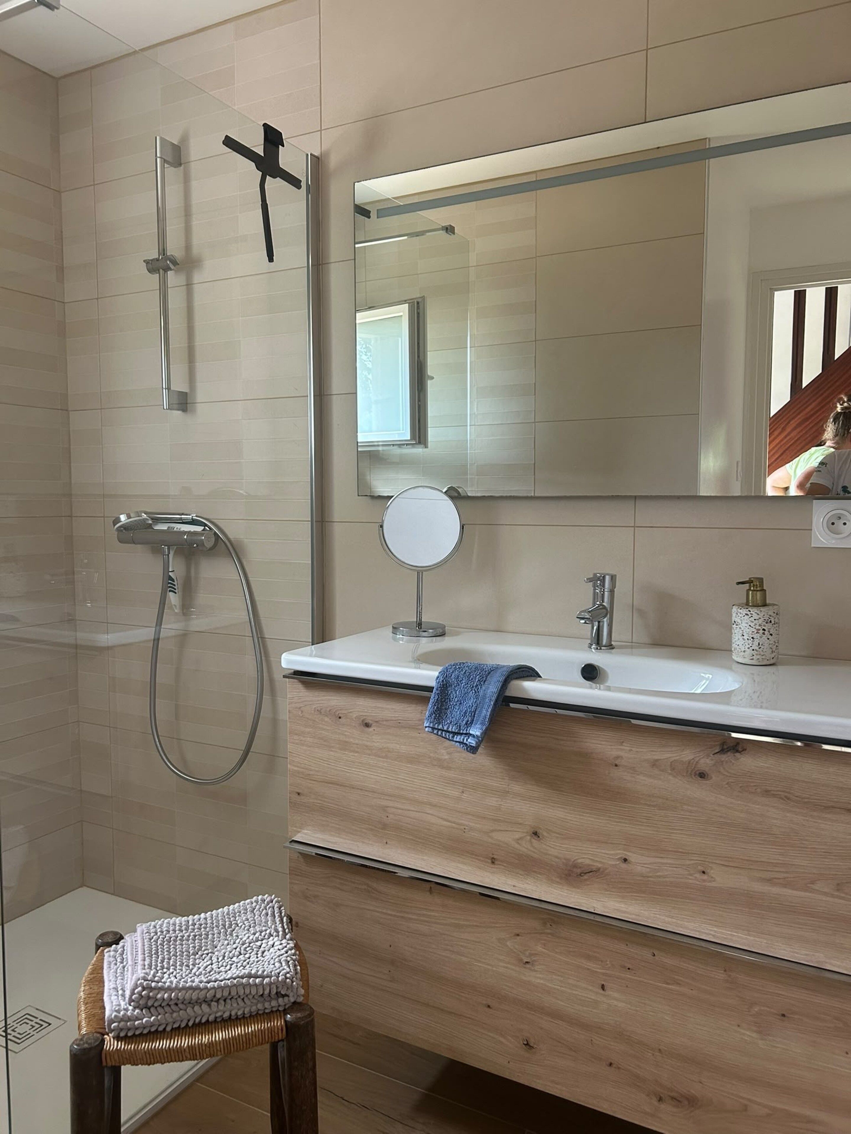 A modern bathroom with a glass shower enclosure, a wooden vanity with a mirror, and a small wicker stool.