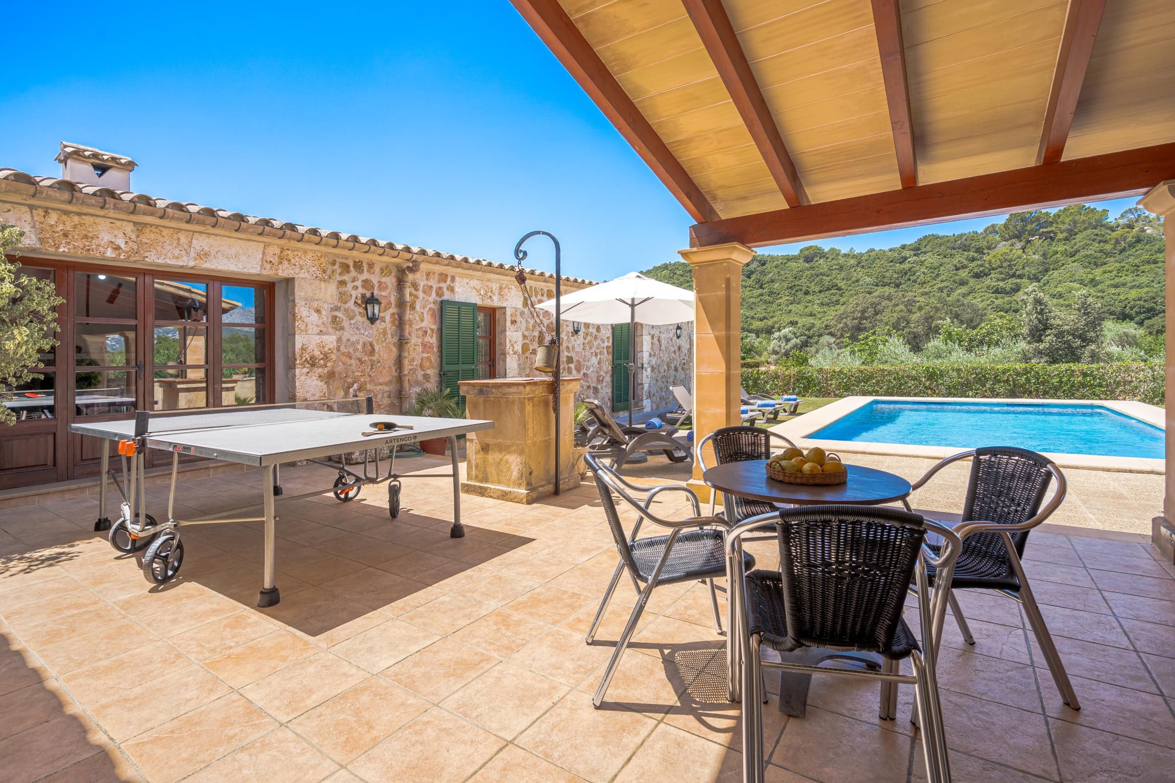 An outdoor patio with a ping pong table, chairs, a table with fruit, a stone wall, and a pool.
