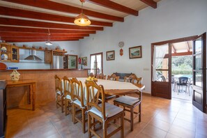 A dining area with wooden chairs and a table, a kitchen with a stove and cabinets, and a window with a view of greenery. - Villa Malagarba - By Dream Villas Pollensa (Pollensa)