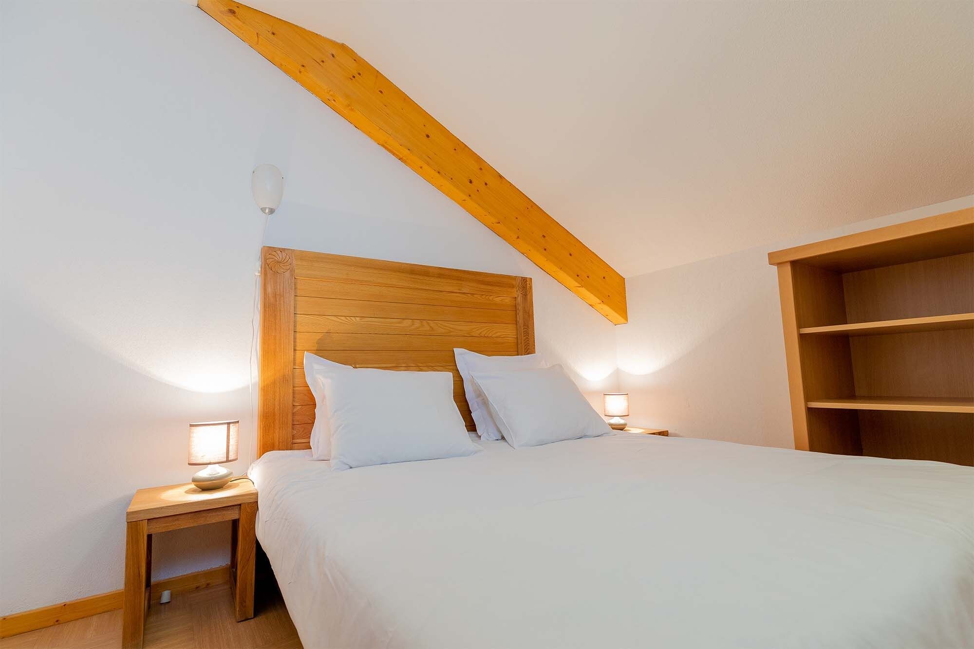 A bedroom with a wooden headboard, a bed with white bedding, two bedside lamps, and a wooden shelf unit.
