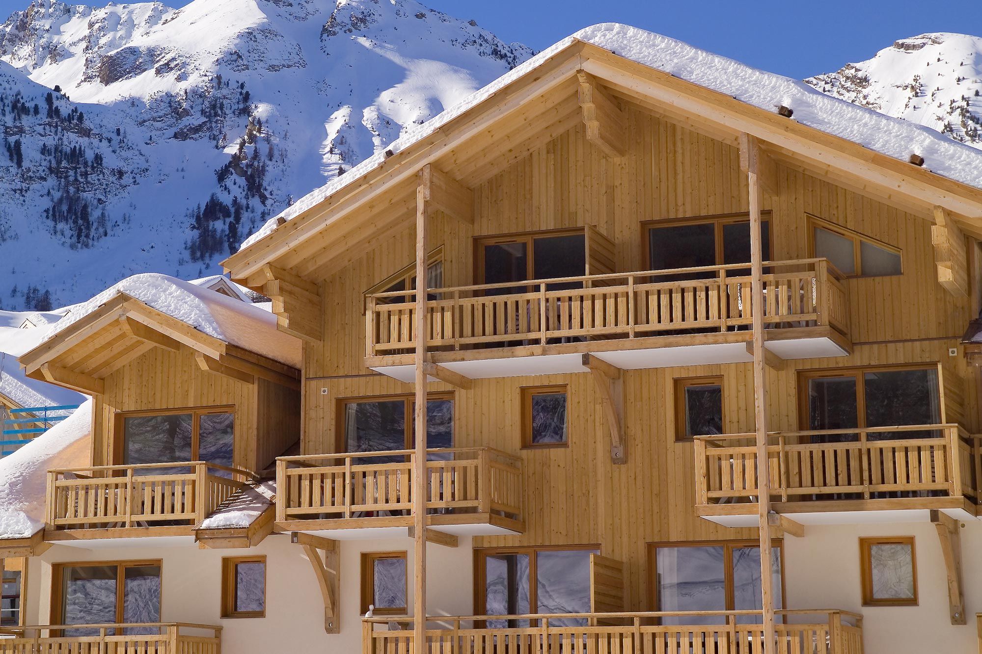 A multi-story wooden chalet with balconies, set against a snowy mountain backdrop.