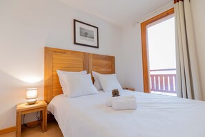 Ein Schlafzimmer mit hölzernem Kopfteil, einem Bett mit weißer Bettwäsche, einem kleinen Nachttisch mit Lampe und einem Balkon, der durch ein offenes Fenster sichtbar ist.