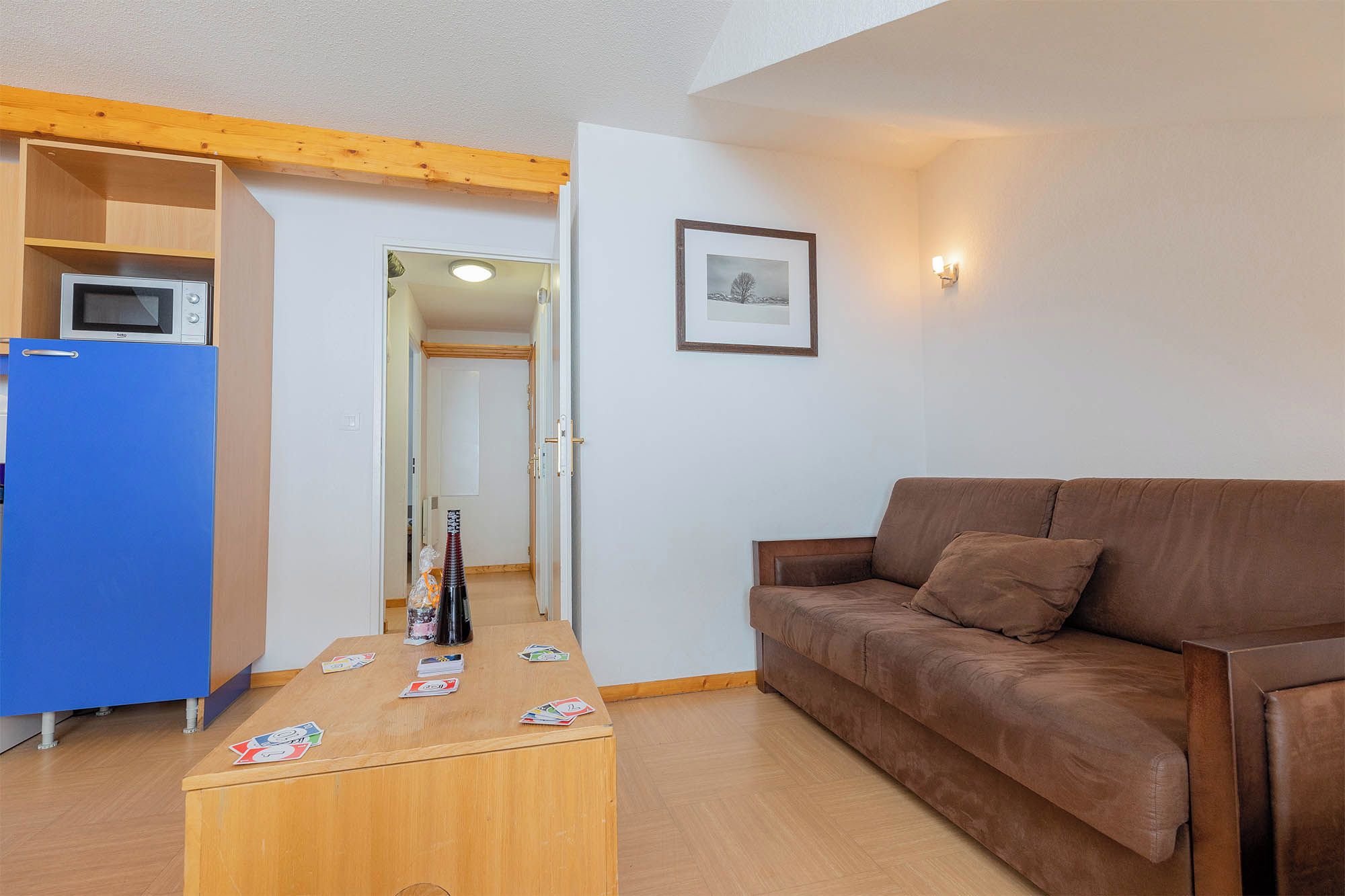Ein Wohnzimmer mit einem braunen Sofa, einem Couchtisch mit Spielkarten, einem blauen Kühlschrank und einem gerahmten Bild an der Wand.