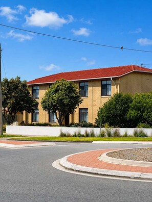 A two-story building with a red-tiled roof, a brick facade, and a landscaped front yard with trees and shrubs. - Driftwood at West Beach - Steps to Sand & Sailing (West Beach)