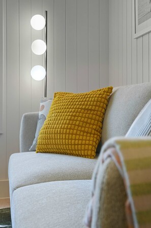A modern living room with a sofa, a yellow textured pillow, and a floor lamp with multiple lights. - Driftwood at West Beach - Steps to Sand & Sailing (West Beach)