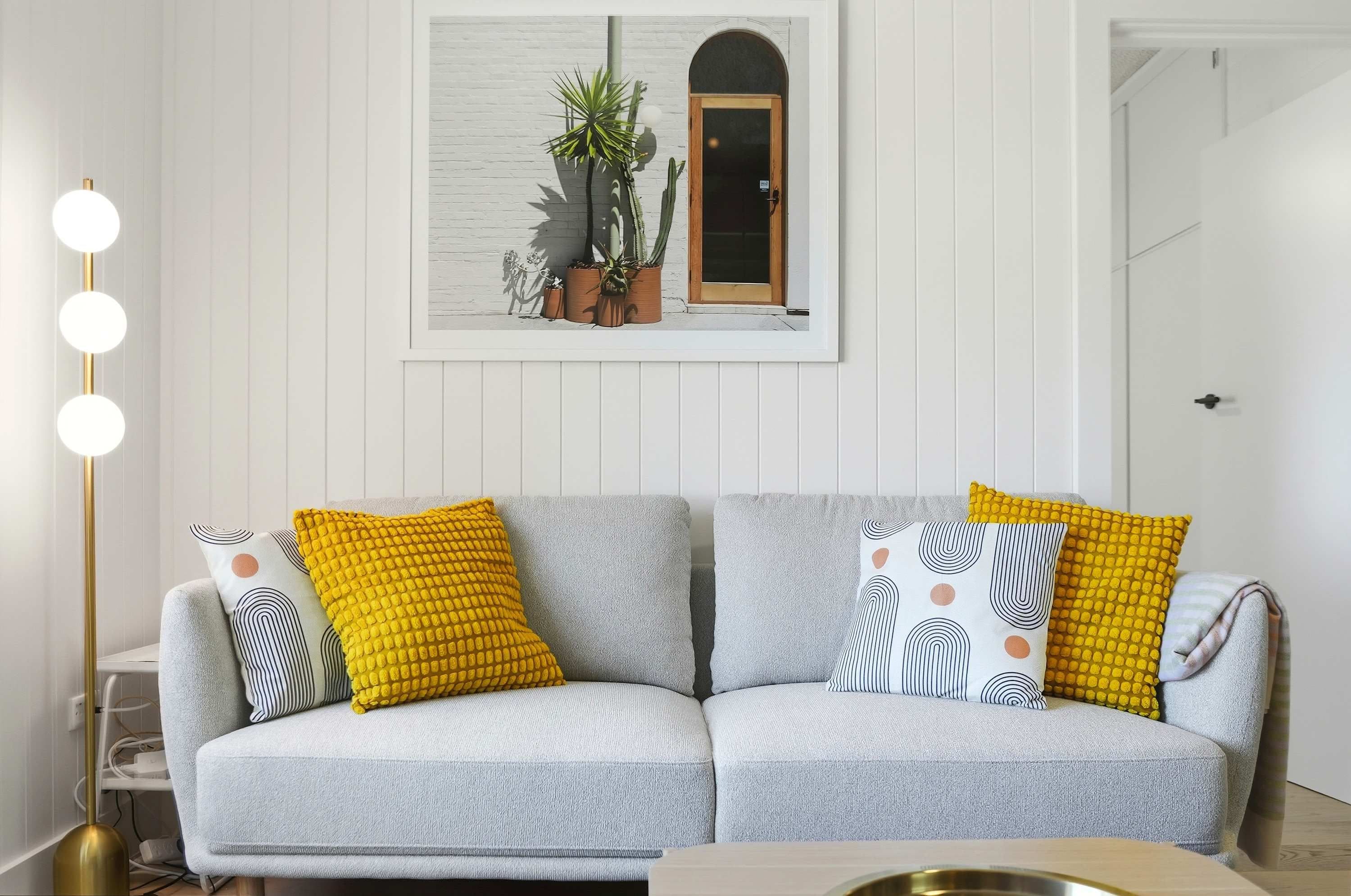 A living room with a grey sofa, yellow and patterned cushions, a floor lamp, and a framed picture of plants on the wall.