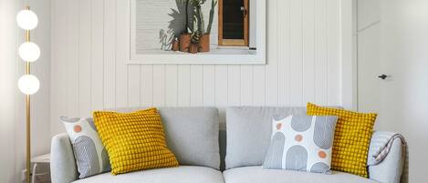 A living room with a grey sofa, yellow and patterned cushions, a floor lamp, and a framed picture of plants on the wall.