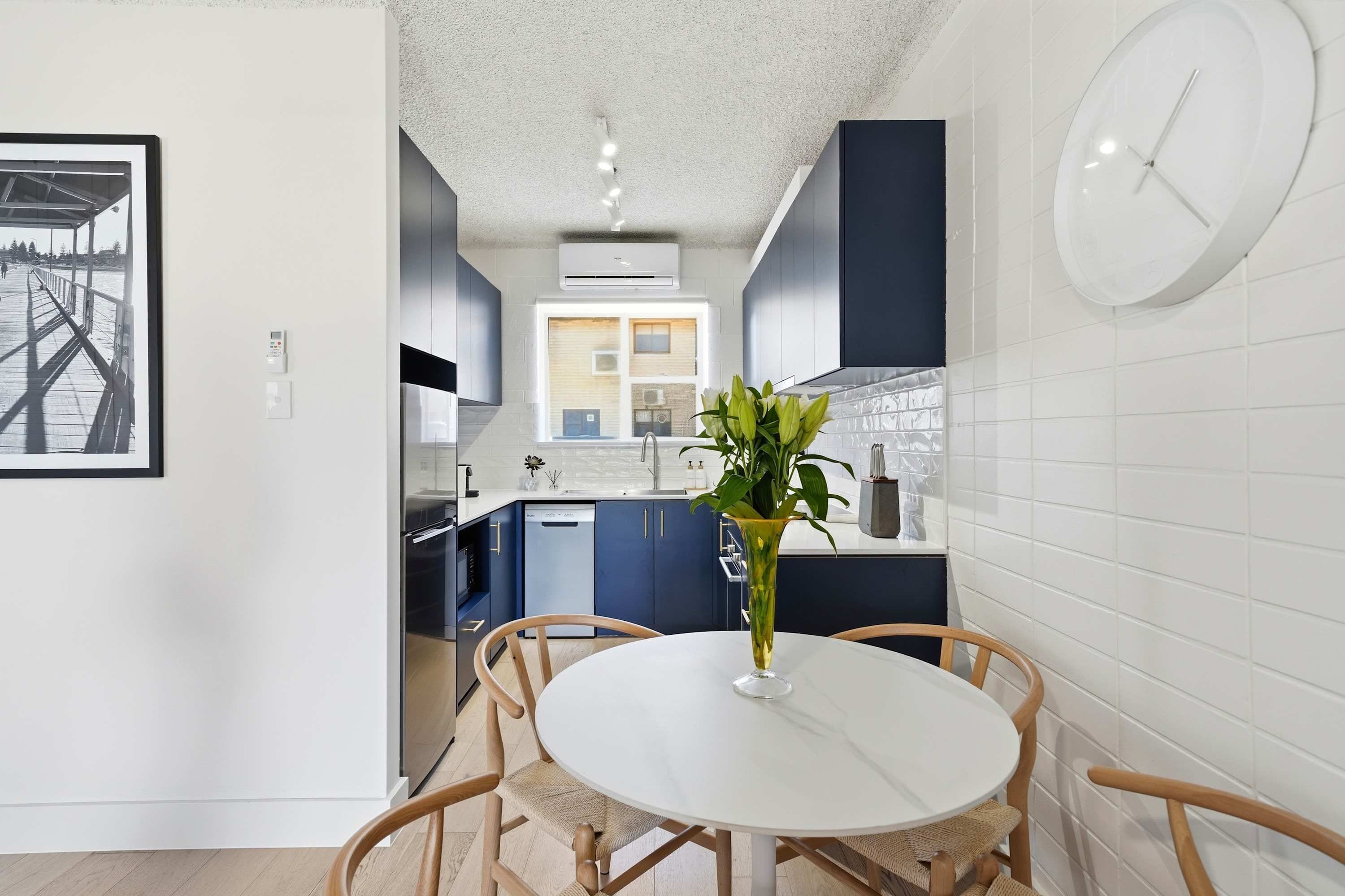 Une cuisine moderne avec des meubles de couleur bleu foncé, un pare-feu en carreaux blancs et une table de salle à manger ronde entourée de chaises en osier.