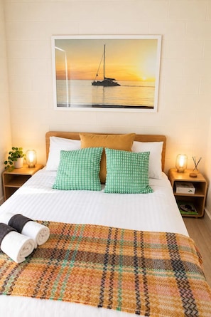 A hotel room with a bed, pillows, a blanket, a painting of a sunset over water, and bedside lamps. - Driftwood at West Beach - Steps to Sand & Sailing (West Beach)