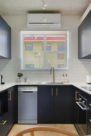 A modern kitchen with a dishwasher, a window with a view of a building, and a white countertop. - Driftwood at West Beach - Steps to Sand & Sailing (West Beach)