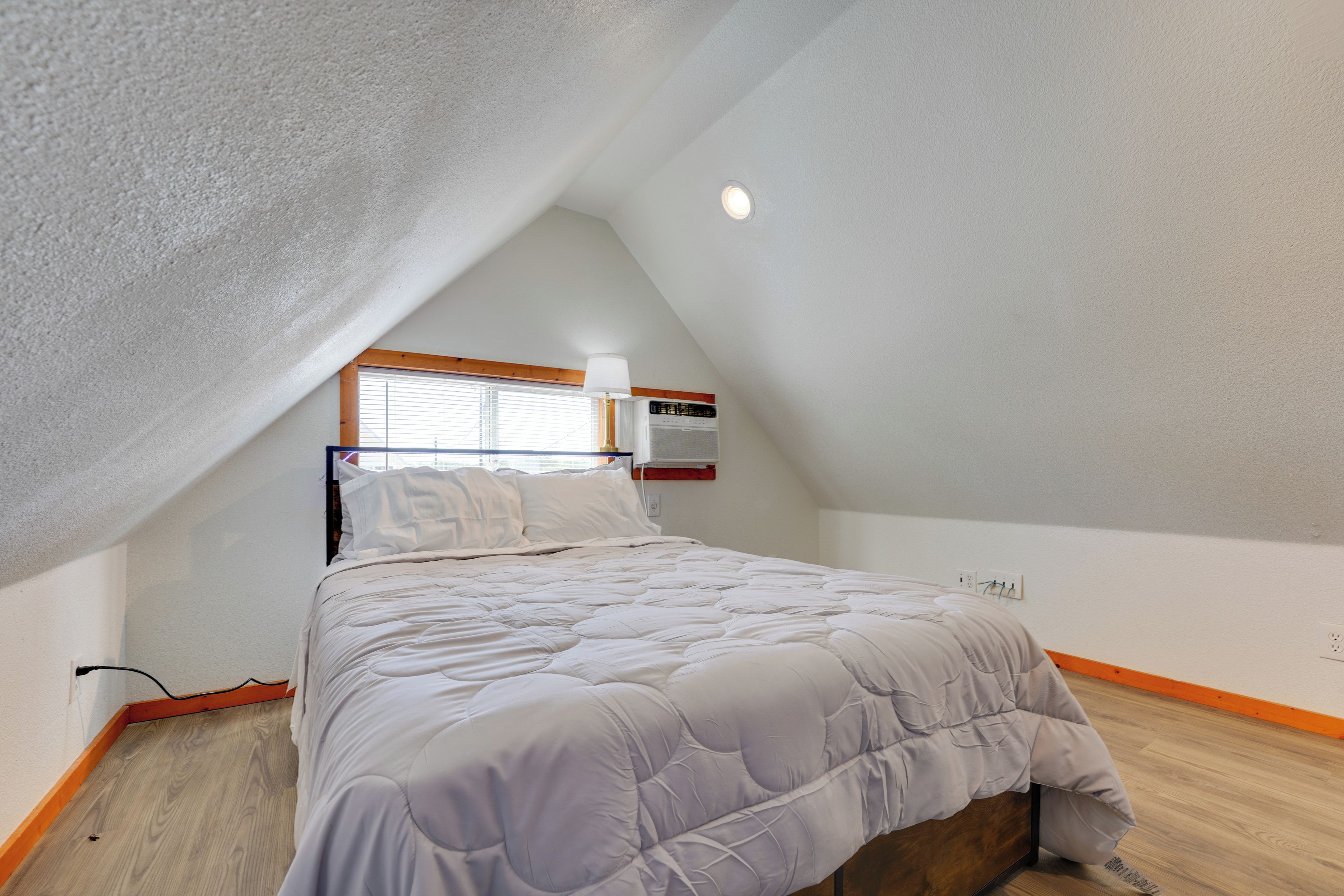 Quarto com teto inclinado, cama com edredom branco, janela com persianas e aquecedor fixado na parede.