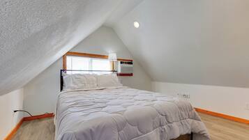 Quarto com teto inclinado, cama com edredom branco, janela com persianas e aquecedor fixado na parede.