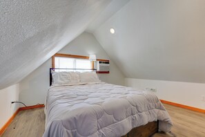 A bedroom with a sloped ceiling, a bed with a white duvet, a window with blinds, and a wall-mounted heater.