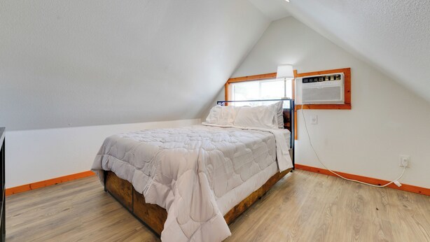 Une chambre avec un plafond en pente, un lit avec un cadre en bois, une fenêtre avec des stores et un climatiseur fixé au mur.