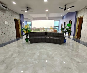 A spacious room with a curved dark-colored sofa, two potted plants, a ceiling fan, and a large window offering a city view. - Deluxe Pratikshya & Pratistha (Balianta)