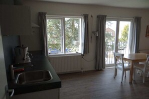 A room with a kitchen area, a dining table with chairs, and a view through a window. - Holiday apartment with 1 bedroom (oostkapelle)