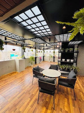 An indoor space with a wooden floor, round tables, and chairs, surrounded by potted plants and a view of buildings. - Shreshtam Signature Serviced Apartment (Hyderabad)