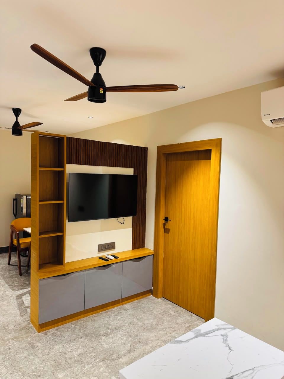 A room with a built-in TV, a wooden door, a ceiling fan, and a small table.