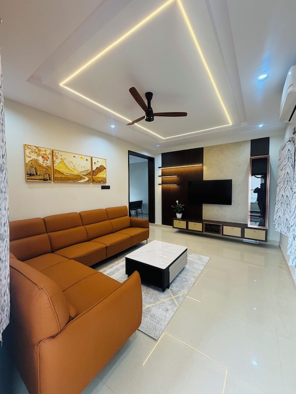 A modern living room with a sectional sofa, a coffee table, a ceiling fan, recessed lighting, and a flat-screen TV mounted on the wall. - Shreshtam Signature Serviced Apartment (Hyderabad)